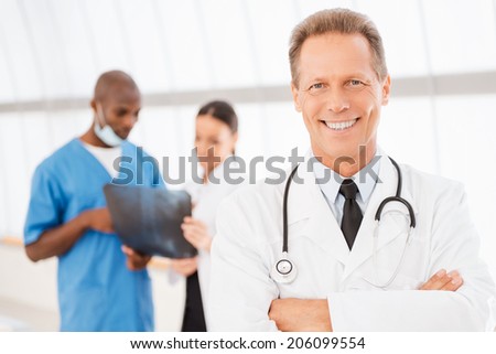 Confident doctor. Cheerful mature doctor keeping arms crossed and smiling while his colleagues discussing x-ray in the background