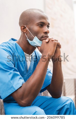 Thoughtful surgeon. Thoughtful young African doctor in blue uniform leaning his face on hands and looking away while sitting at the chair in corridor