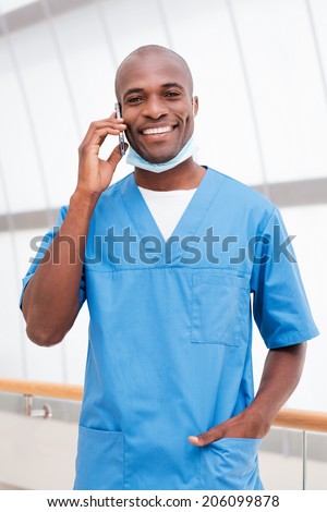 Doctor on the phone. Cheerful young African doctor in blue uniform talking on the mobile phone and looking at camera