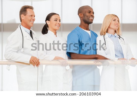 Confident team of medical experts. Low angle view of four confident doctors standing close to each other and looking away while leaning at the handrail