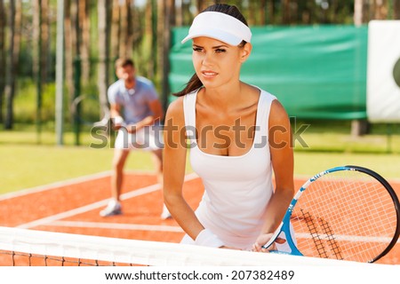 They play like a team. Beautiful young woman holding tennis racket and looking away while standing on tennis court and with man in the background