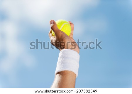 Tennis ball in his hand. Close-up of male hand in wristband holding tennis ball against blue sky