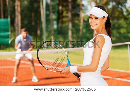 Feeling confident of winning. Beautiful young woman holding tennis racket and looking over shoulder with smile while man in sports clothing standing in the background