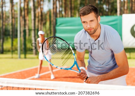 Concentrated on game. Handsome young man holding tennis racket and looking away while standing on tennis court and with woman in the background