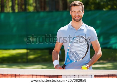 Great day to play! Cheerful young man in polo shirt holding tennis racket and ball while standing on tennis court