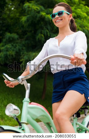 Riding her new bike in park. Low angle view of attractive young smiling woman in sunglasses riding her vintage bicycle in park