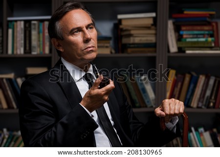 Thoughtful gentleman. Confident mature man in formalwear holding pipe and cane while sitting against bookshelf