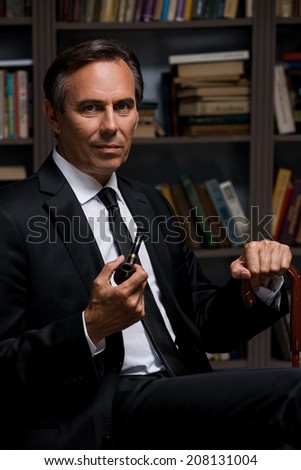 Confident gentleman. Confident mature man in formalwear holding pipe and cane while sitting against bookshelf