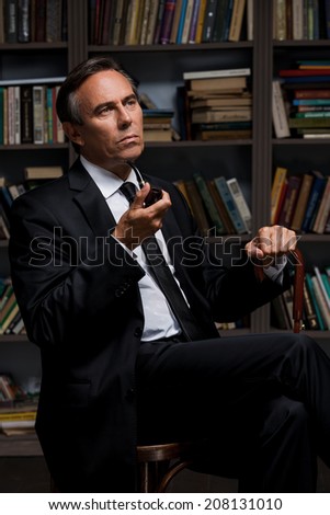 Gentleman with pipe. Thoughtful mature man in formalwear holding pipe and cane while sitting against bookshelf