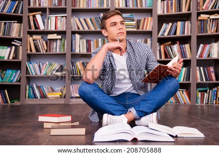 Preparing to his final exams. Thoughtful young man holding note pad and touching his chin with pen while sitting against bookshelf