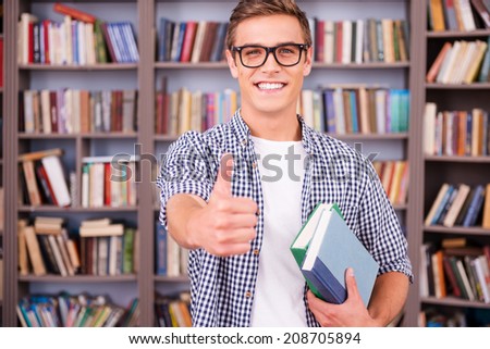 He loves studying. Handsome young man holding books and showing his thumb up while standing in library