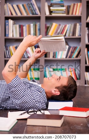 Happy bookworm. Side view of happy young man reading book while lying on the floor against bookshelf