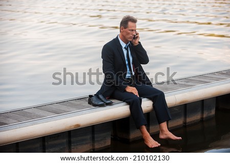 Bad business news. Depressed mature businessman talking on the mobile while sitting barefoot at the quayside