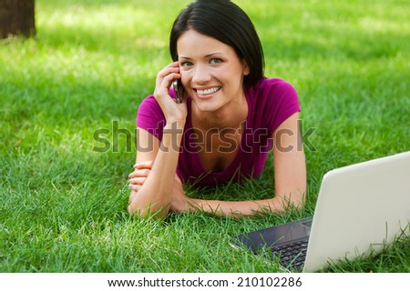 Technologies making life easier. Attractive young woman talking on the mobile phone and smiling while lying in grass with laptop laying near her