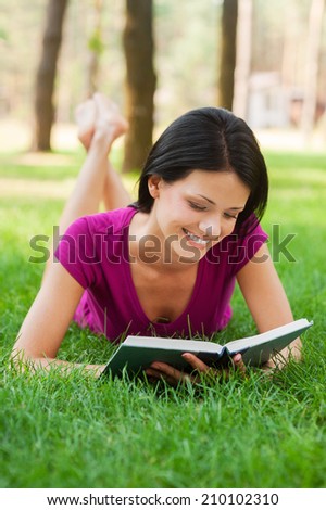 Student in nature. Attractive young woman reading book and smiling while lying in grass
