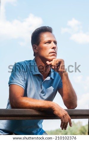 Waiting for inspiration. Thoughtful mature man holding hand on chin and looking away while leaning at the wooden fence