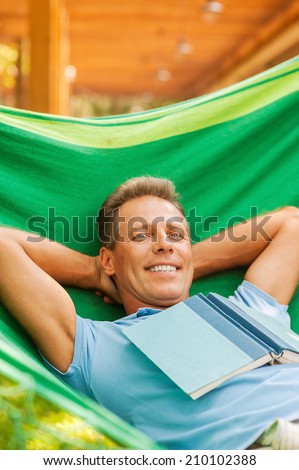 So good to do nothing. Happy mature man sleeping while lying in hammock with book laying on his torso