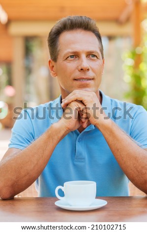 Inspired with cup of fresh coffee. Thoughtful mature man holding hands on chin while sitting in sidewalk cafe with coffee cup on the table