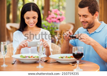 Taking pictures of food. Beautiful young loving couple taking pictures of their food and smiling while relaxing in outdoors restaurant together