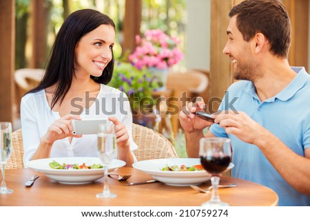 Capturing their time together. Happy young loving couple taking pictures of their food and smiling while relaxing in outdoors restaurant together