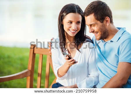 Look at this picture! Beautiful young loving couple sitting on the bench while woman showing mobile phone to her boyfriend and smiling