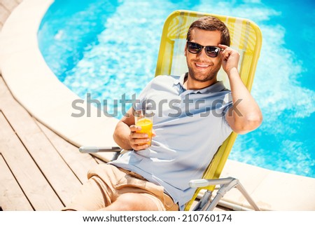 This is the life! Top view of cheerful young man in polo shirt sitting at the deck chair and holding glass with orange juice