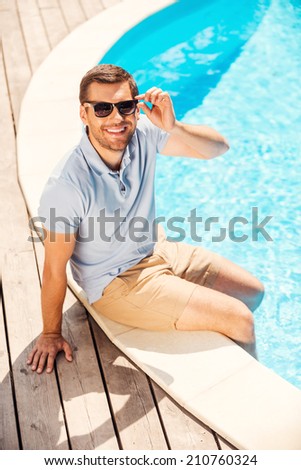 Summer time fun. Top view of cheerful young man in polo shirt sitting by the pool and adjusting his sunglasses