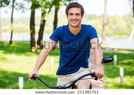 Enjoying his park ride. Cheerful young man riding bicycle in park and smiling at camera