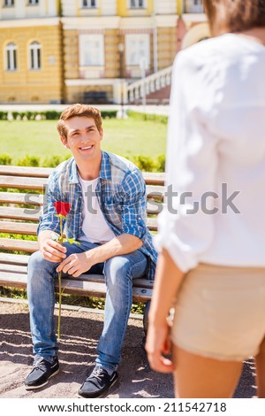 Finally meet! Happy young man sitting on the bench and holding single rose with woman on foreground
