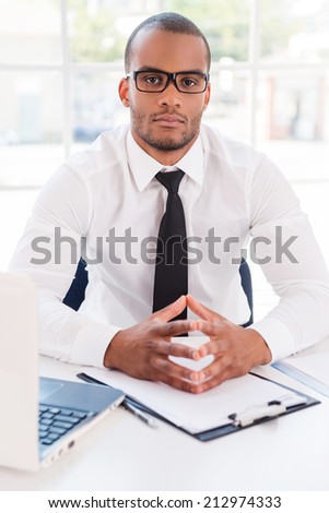 Confident businessman. Confident young African man in shirt and tie looking at camera while sitting at his working place