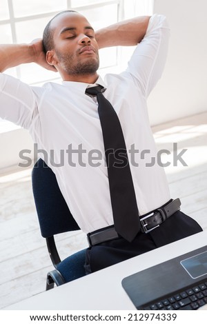 Total relaxation. Relaxed young African man in shirt and tie holding hands behind head while sitting at his working place