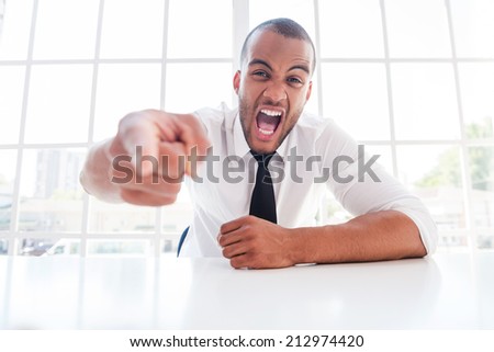 Furious boss. Furious young African man in shirt and tie pointing you and shouting while sitting at his working place