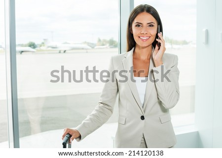 Waiting for flight. Beautiful young businesswoman in formalwear talking on the mobile phone and smiling while standing in airport with airplanes in the background