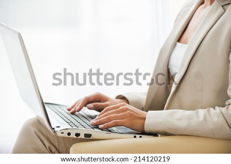 Working on laptop. Side view of young businesswoman in suit working on laptop while sitting at the chair