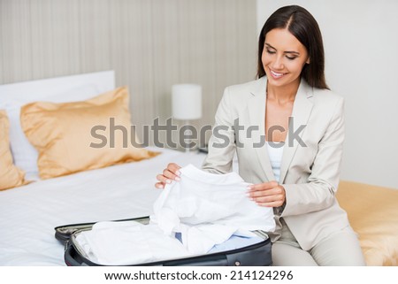 Unpacking her suitcase. Beautiful young businesswoman in suit unpacking her suitcase and smiling while sitting on the bed in hotel room