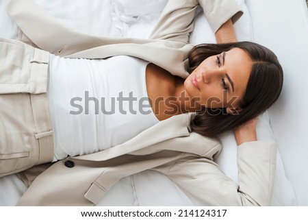 Tired after hard working day. Top view of beautiful young businesswoman in suit holding hands behind head and keeping eyes closed while lying in bed at the hotel room
