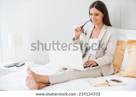 Working in hotel room. Beautiful young businesswoman in suit working on laptop and smiling while sitting in bed at the hotel room