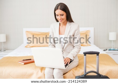 Staying connected anytime. Beautiful young businesswoman in suit working on laptop and smiling while sitting on the bed in hotel room