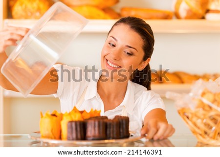 The freshest bakery for our customers. Beautiful young woman in apron carrying plate with fresh cookies and smiling while standing in bakery shop