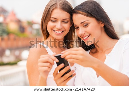 Look at this! Two beautiful young women looking at mobile phone and smiling while standing outdoors together