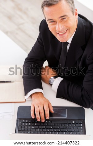 Businessman at work. Top view of happy mature man in formalwear working on laptop and smiling while sitting at his working place