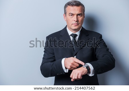 Time is money. Confident mature man in formalwear pointing his watch and looking at camera while standing against grey background