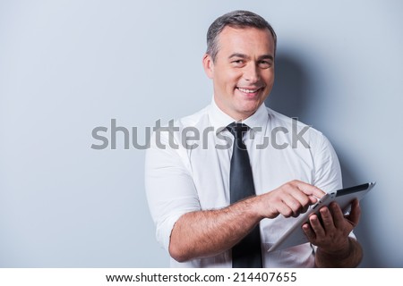 Examining his brand new tablet. Confident mature man in shirt and tie working on digital tablet and smiling while standing against grey background