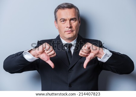 Bad news for you. Confident mature man in formalwear showing his thumbs down and looking at camera while standing against grey background