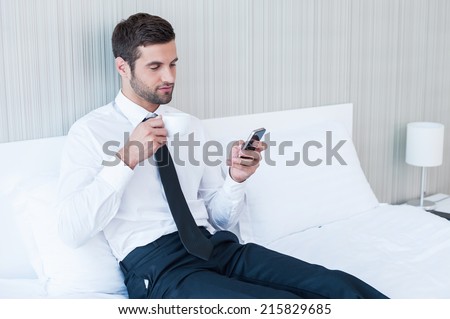 Typing business message. Confident young man in shirt and tie drinking coffee and holding mobile phone while lying in bed at the hotel room
