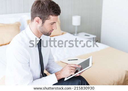 Working in hotel room. Confident young businessman in shirt and tie working on digital tablet while sitting on the bed in hotel room