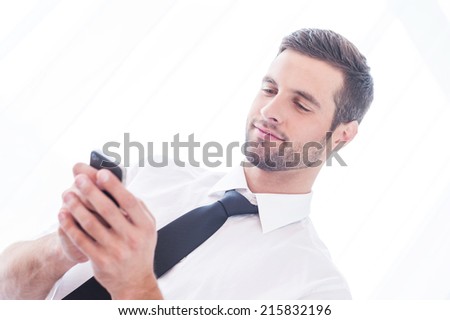 Typing business message. Low angle view of handsome young man in shirt and tie typing message on the mobile phone and smiling