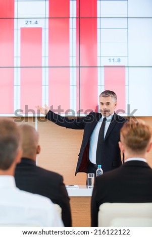 Talking about company achievements. Confident mature man in formalwear pointing projection screen with graph on it while making presentation in conference hall with people on the foreground