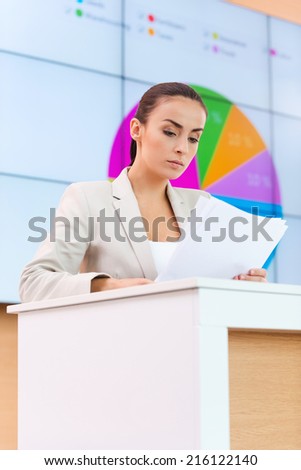 Confident public speaker. Confident young woman in formalwear standing at the tribune and looking at paper while making a presentation with projection screen in the background