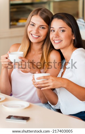 Friends in coffee shop. Two attractive young women holding coffee cups and smiling while sitting in coffee shop together